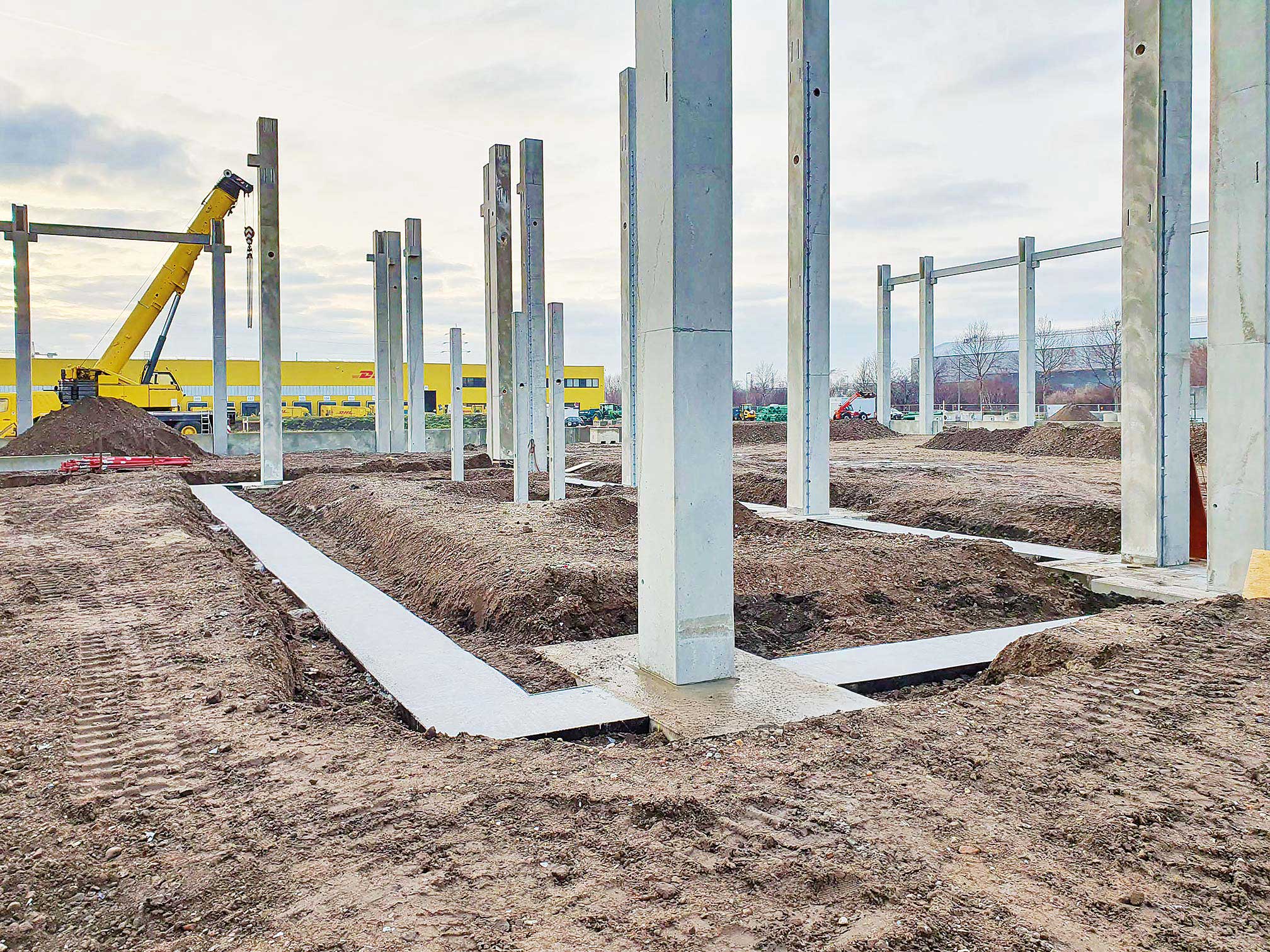 Baustelle mit Betonpfeilern und einem gelben Kran vor einem gelben Gebäude bei bewölktem Himmel.