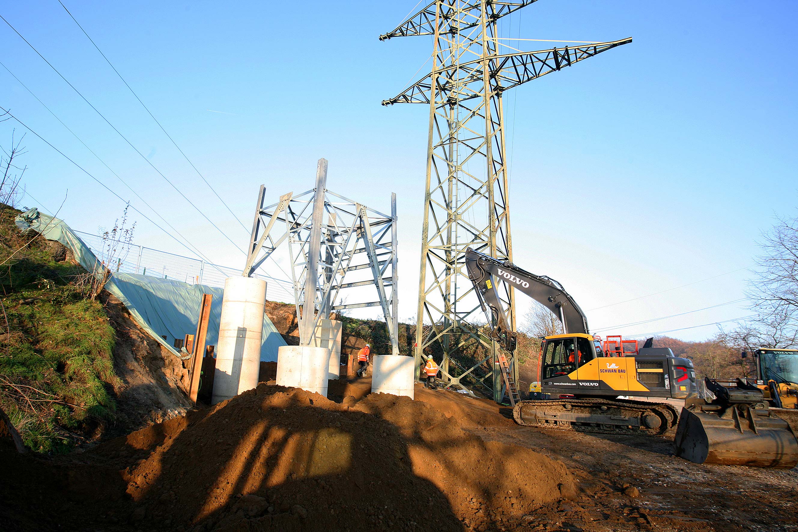 Baustelle mit Volvo-Bagger, Bauarbeitern und Stahlkonstruktion unter Hochspannungsmasten bei klarem Himmel.