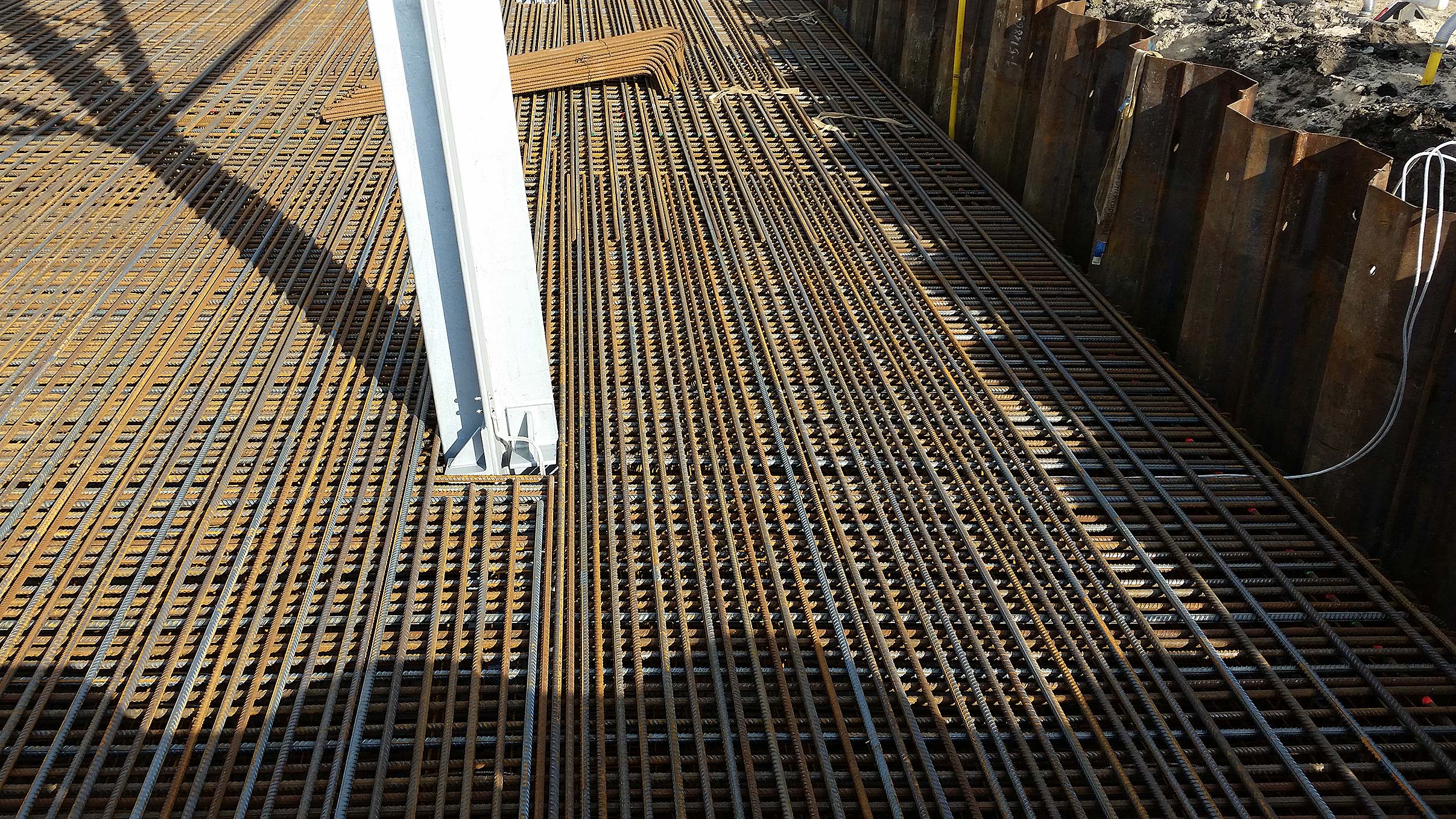 Stahlbewehrung auf einer Baustelle, vorbereitet für Beton, mit einem weißen Stahlträger und Stahlbohlen im Hintergrund.